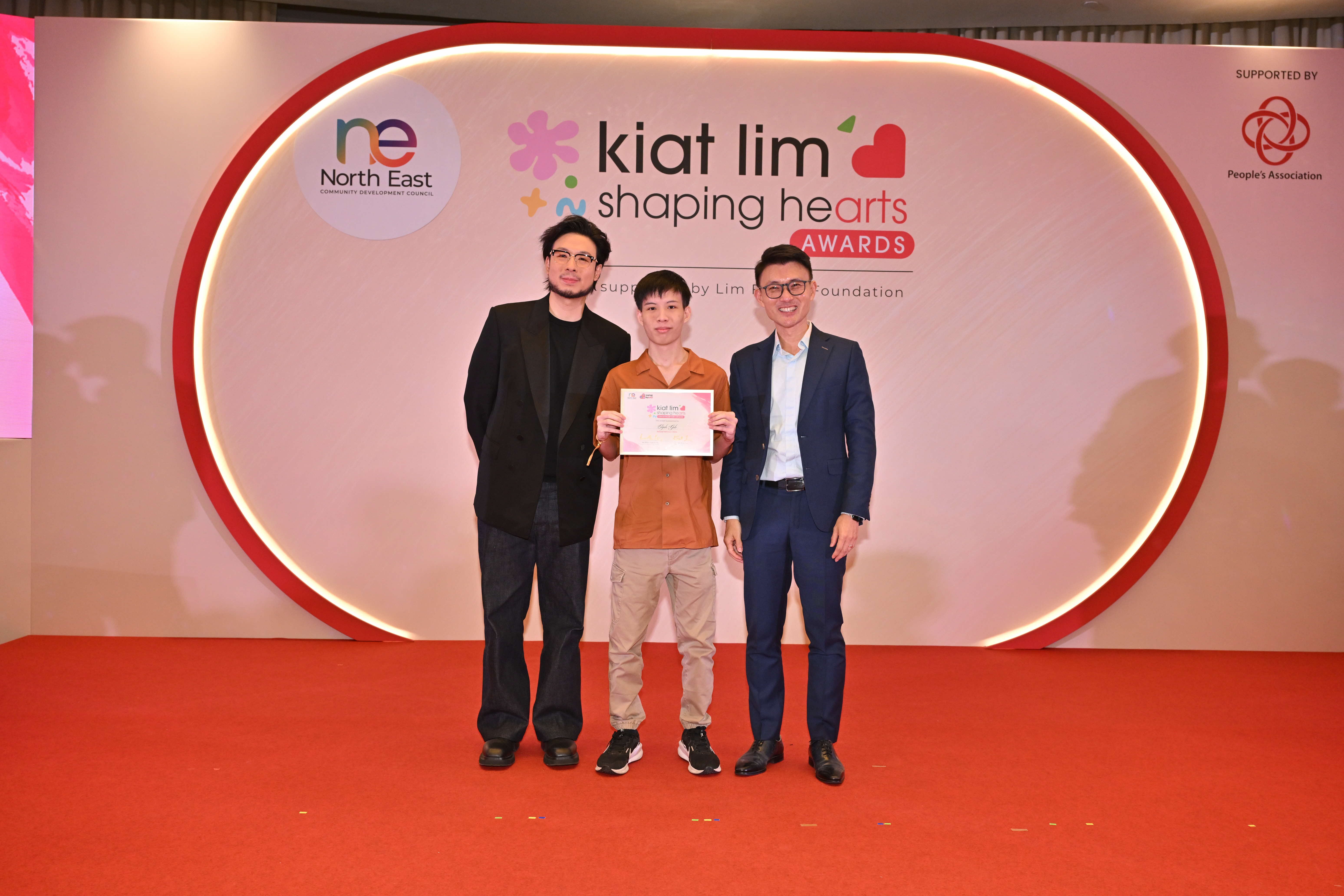 Award recipient standing on stage with the presenter, holding their certificate and posing for a group photo during the award ceremony, with a backdrop reading ‘Kiat Lim-Shaping Hearts Awards'.